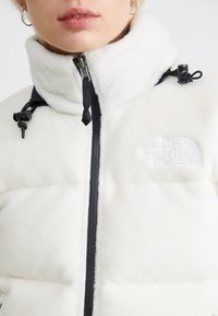 Biely fleece kabát s vysokým golierom, čiernym zipsom a šnúrkovými detailmi. Na hrudi je vyšívané logo. Mäkká textúra.