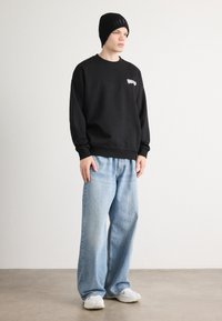 Felpa nera con logo sul petto, abbinata a jeans baggy azzurri chiari e sneakers bianche. Il modello indossa un berretto a coste nero.