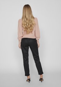 Kobieta z długimi falującymi blond włosami nosi różowy sweter, czarne spodnie z podwiniętymi nogawkami oraz czarne sandały na obcasie, stojąc na jednolitym jasnym tle.