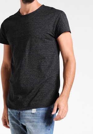 Uomo che indossa una semplice t-shirt grigia scura a maniche corte e jeans scoloriti azzurri chiari, in piedi di fronte a uno sfondo bianco uniforme.