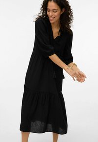 Svart klänning i medellängd med trekvartsärmar, med ett lager på lager-design, tillverkad av lättviktigt tyg, med v-hals och fransade snören.