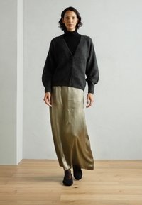 Mörkgrå stickad cardigan med v-ringning och knappar, bärs över en lång sidenkjol i guld. Svarta skor fulländar outfiten.