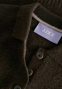 Cardigan en laine marron avec col replié, trois boutons, et une étiquette lavande avec "JJXX". Tissu doux et texturé avec une apparence confortable.