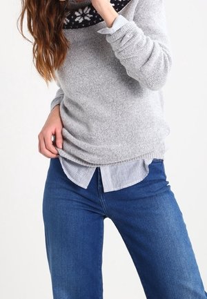 Vrouw draagt een blauwe spijkerbroek, een gestreept shirt en een lichtgrijze trui met een zwart-wit sneeuwvlokpatroon bij de kraag.