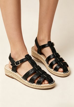 Pieds portant des sandales en cuir texturé noir avec semelles plateformes en jute tissé et brides ajustables à la cheville sur un fond clair.