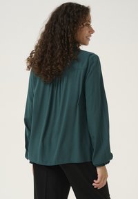 Blusa lungo maniche color teal con dettagli arricciati sulle spalle e orlo arrotondato. Il tessuto appare morbido con una texture liscia.