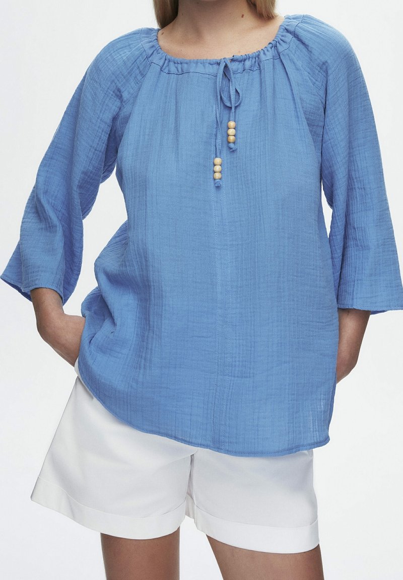 Blusa azzurro chiaro, testurizzata, con scollo rotondo, chiusura con lacci regolabile e maniche a campana tre quarti. Presenta dettagli con perline di legno.