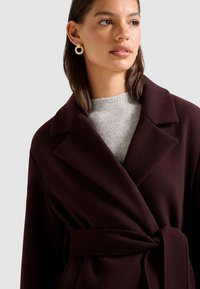 Abrigo burdeos cruzado con cuello con muesca, cintura con cinturón, textura suave y suéter gris debajo. Modelo lleva pendientes circulares de oro.