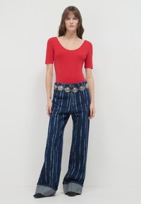 Donna che indossa un top rosso a scollo rotondo, jeans a gamba larga blu scuro a righe con grandi risvolti, e una cintura di monete d'argento, in piedi contro un muro bianco.