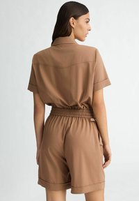Jumpsuit marrone a maniche corte con colletto, vita elastica e tessuto liscio dalla vestibilità comoda. Il design sul retro presenta un dettaglio in yoke.