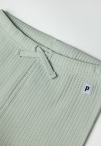 Lichtgroene geribbelde stof met een tailleband met trekkoord en een klein label met een "P"-logo. De gestructureerde oppervlakte geeft dimensie.