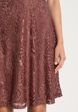 Main de femme reposant à côté d'une jupe en dentelle mauve mi-longue avec des motifs floraux et un ourlet festonné.