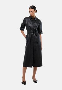 Robe chemise en simili cuir noir avec des manches longues, une taille ceinturée, une fermeture à boutons sur le devant et une longueur au genou. Texture lisse avec une finition brillante.