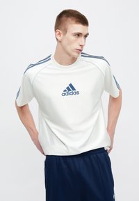 Biele športové tričko s modrými pruhmi na ramenách, s tmavomodrým logom Adidas na prednej strane. Krátke rukávy, okrúhly výstrih.