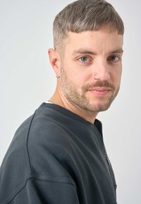 Dark gray zip-up sweatshirt with a round neckline, textured fabric, and visible stitching details. Short, straight-cut hair and facial stubble.