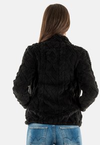 Chaqueta de pelo sintético negra con un patrón de diamantes texturizado, que presenta un cuello y mangas largas, llevada sobre unos vaqueros de mezclilla azules.