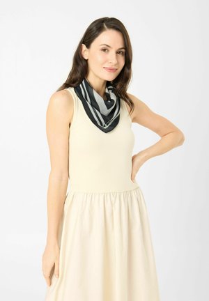 Frau mit dunklen Haaren, die ein ärmelloses beiges Kleid und einen schwarz-grau gestreiften Schal trägt, steht mit einer Hand in der Hüfte.