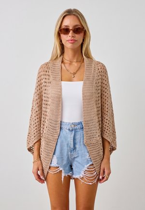 Vrouw draagt bruine gehaakte open cardigan, wit topje, versleten spijkershorts, zonnebril en gouden sieraden, staand voor een effen achtergrond.