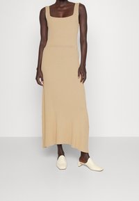 Beige Strick-Maxi-Kleid mit quadratischem Ausschnitt und gerippter Textur. Kombiniert mit weißen, hohen Mules, die eine elegante Silhouette betonen.