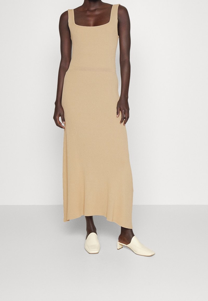 Beige Strick-Maxi-Kleid mit quadratischem Ausschnitt und gerippter Textur. Kombiniert mit weißen, hohen Mules, die eine elegante Silhouette betonen.