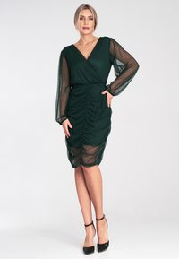 Ciemnozielona sukienka o kroju kopertowym, z przezroczystymi długimi rękawami, marszczoną teksturą w talii oraz długości do kolan. Założona z czarnymi szpilkami.