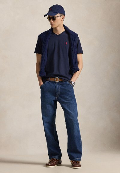 Navy majica s V-izrezom, mornarsko plavi džemper pleten mrežasto prebačen preko ramena, plave traperice, smeđe cipele za čamce i tamne sunčane naočale.