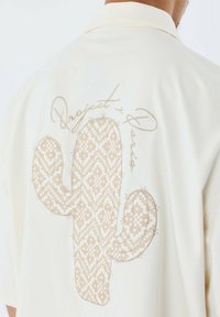 Camisa de manga corta color crema con un gran diseño de cactus beige en la parte trasera, que presenta patrones geométricos y detalles de costura fina.