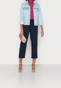Chaqueta de mezclilla azul claro, top púrpura de cuello alto, pantalones navy recortados, tacones nude con tiras y un bolso metálico. Silueta limpia y estructurada.