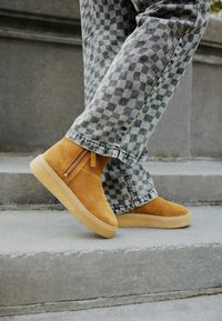 Botines de ante color beige con puntera redondeada, que cuentan con una cremallera lateral y una ligera suela de goma, combinados con pantalones de mezclilla gris a rayas.