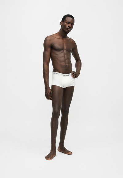 Λεπτός μαύρος ανδρικός μοντέλο στέκεται ξυπόλυτος με λευκά μποξεράκια Off-White, ένα χέρι στη μέση, πάνω σε απλό ανοιχτό γκρι φόντο.