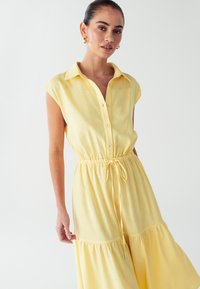 Geltonas marškinėlių suknelė su apykakle, trumpomis rankovėmis, mygtukais priekyje, elastinga juosmens juosta ir sluoksniuota sijonėliais. Pagaminta iš lengvos, minkštos medžiagos.