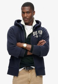 Felpa con zip di colore blu scuro con un logo, indossata sopra una polo verde, pantaloni beige e un orologio giallo al polso, con uno sfondo neutro.