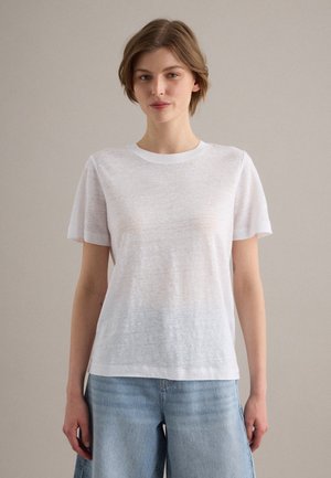 Joven mujer con pelo corto y castaño, vestida con una camiseta blanca lisa de manga corta y vaqueros de mezclilla azul claro, de pie frente a un fondo neutro.