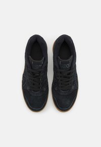 Sapatilhas de camurça pretas com um cabedal texturizado, apresentando um colarinho acolchoado, atacadores pretos e uma sola de borracha natural. Costuras detalhadas em destaque.