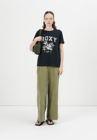 Zwarte t-shirt met witte bloemen- en tekstprint, gecombineerd met olijfgroene wijde corduroypanten. Model houdt een kleine groene tas vast.