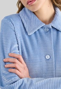 Mujer que lleva un abrigo abotonado de textura azul claro con un cuello ancho, cruzando los brazos, con los labios ligeramente entreabiertos, sobre un fondo neutral.