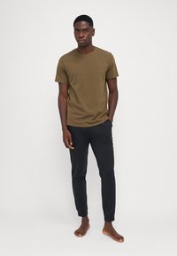 Olivově zelené bavlněné tričko s krátkým rukávem, spárované s černými tapered jogger kalhotami. Model stojí bosý na jednoduchém bílém pozadí.