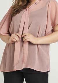 Blouse rose clair avec un col à nouer, texture semi-transparente et manches courtes volantées, superposée à un haut noir ajusté.
