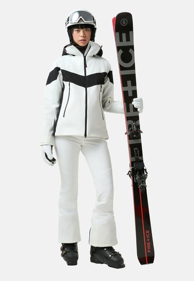 Λευκό και μαύρο μπουφάν για σκι με σχέδιο σε σχήμα βέλους, σε συνδυασμό με λευκό παντελόνι για σκι και μαύρα παπούτσια για σκι· κρατώντας κόκκινα και μαύρα σκι με δεσίματα.