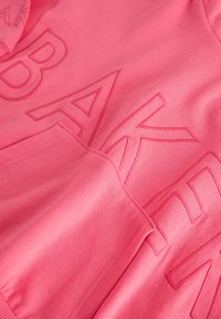 Nahaufnahme von rosa Stoff mit erhabenen, bestickten Buchstaben, die einen Teil des Wortes „BAKER“ bilden, und einer vorderen Kängurutasche.
