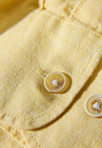 Geel textiel met een fijne textuur, voorzien van twee ronde beige knopen. De stiksels zijn zichtbaar, wat de ontwerpdetails benadrukt.