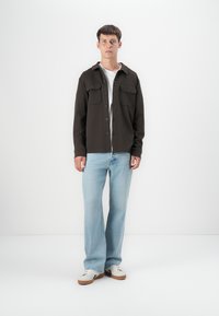 Chaqueta de camisa marrón oscuro con dos bolsillos en el pecho, combinada con jeans azul claro y zapatillas blancas. Tejido suave, corte relajado.