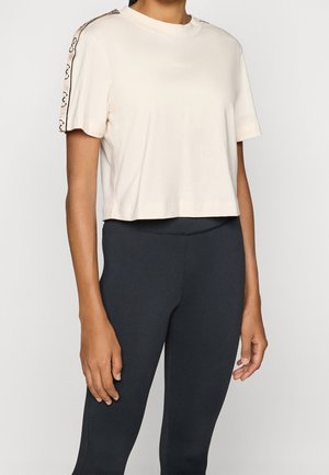 Vrouw draagt een beige korte mouw crop top met logo strepen op de mouwen en een zwarte legging met hoge taille, tegen een effen achtergrond.