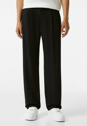 Homme portant un pantalon noir à jambes droites et des baskets blanches, debout les mains détendues le long du corps sur fond blanc.