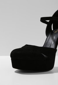 Chaussure à talon haut plateforme en daim noir avec bride à la cheville, bout arrondi et semelle intérieure en cuir lisse sur une surface claire.