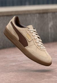 Beige mokkasukot kengät ruskeilla nahka-akseleilla, joissa on kumipohja, valkoiset nauhat ja kultainen logo sivussa. Klassinen retrodesign.