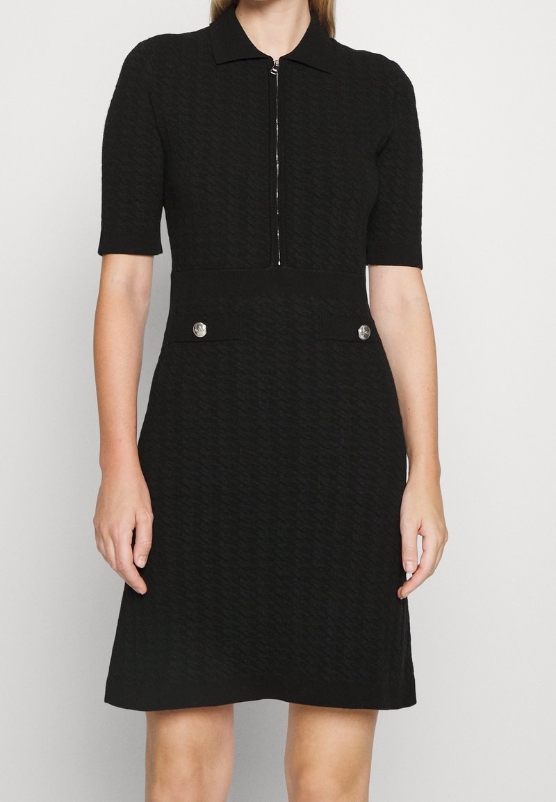 Robe noire texturée longueur genoux avec manches courtes, col, fermeture éclair devant et deux poches décoratives à boutons argentés à la taille.