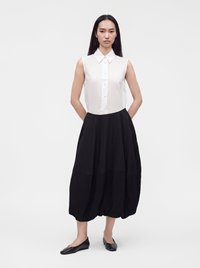 Femme aux longs cheveux foncés portant une chemise blanche sans manches boutonnée, une jupe noire ample en forme de bulle et des chaussures plates noires, debout les mains derrière le dos.