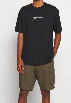 Black cotton t-shirt with short sleeves, featuring a white cursive logo on the front. Paired with olive green cargo shorts.