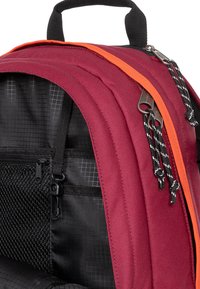 Maroonin reppu oransseilla korostuksilla, mustilla vetoketjuilla ja hihnoilla. Tekstuuroitu kangas, jossa on verkko-osa ja kestävä vetoketju.